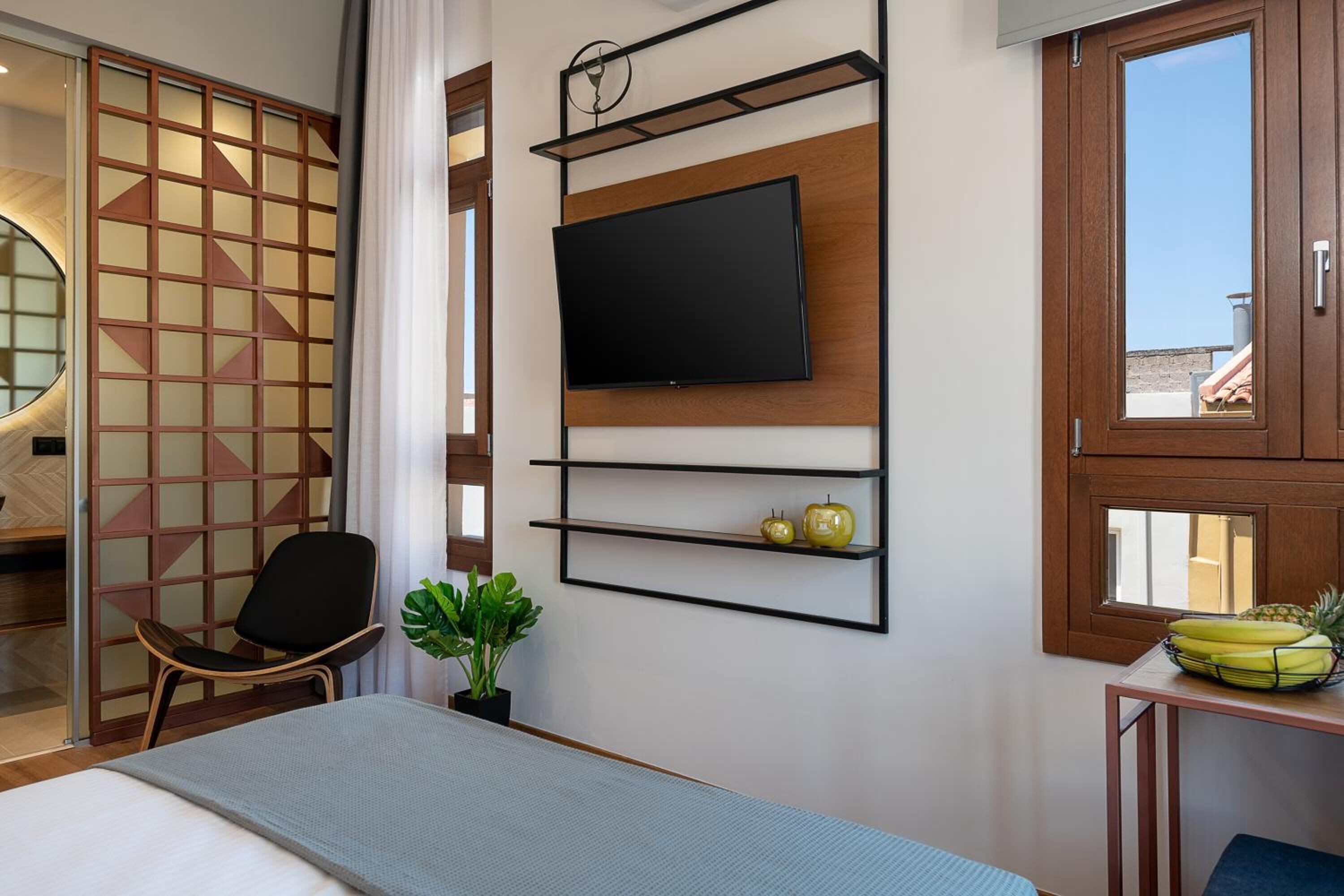 A hotel room with a bed, a TV mounted on the wall, a chair, a small table with fruit, and a window with a view.