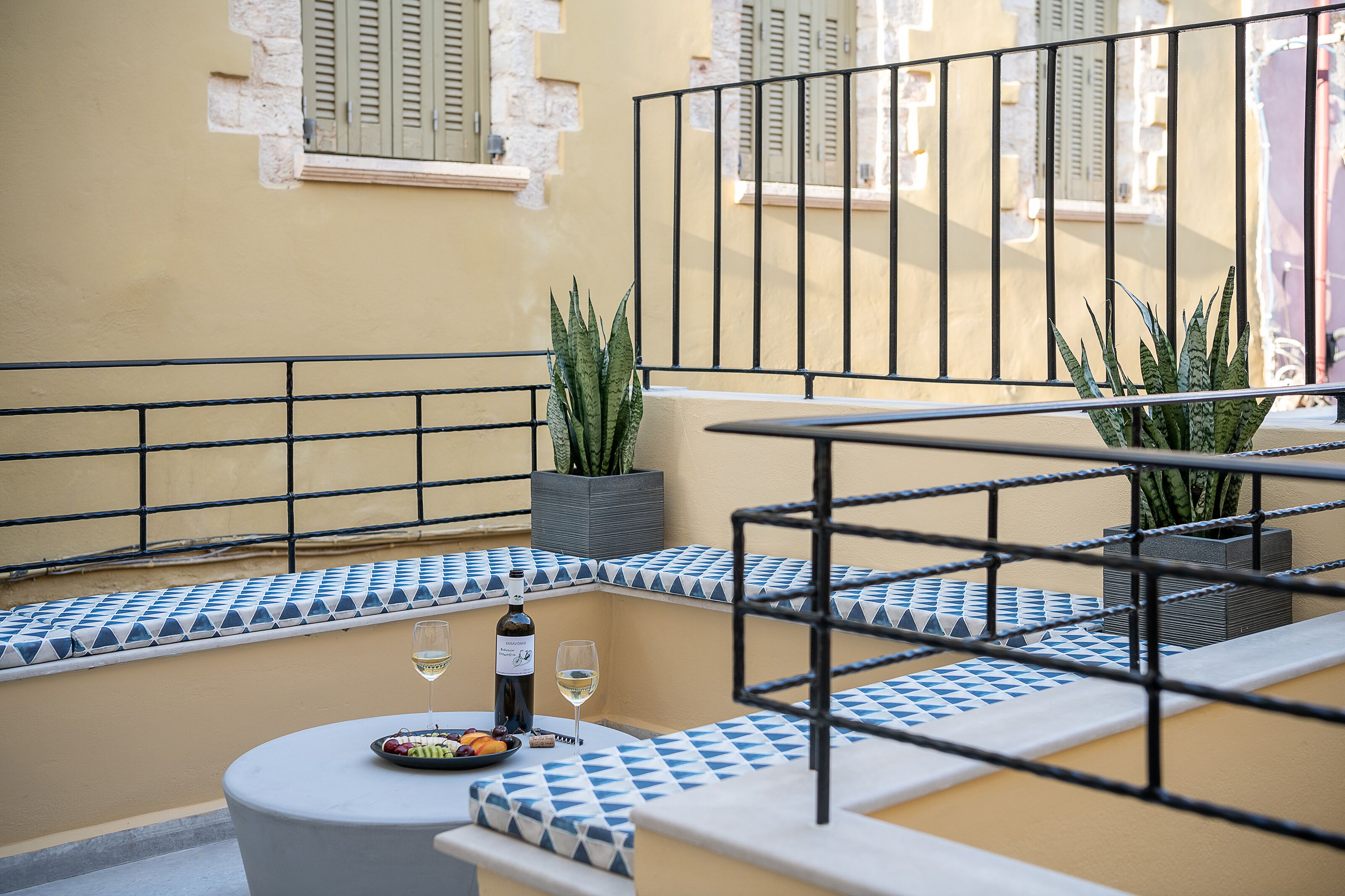 A balcony with a table set with a bottle of wine, a glass, and a plate of food. There are potted plants and a railing.