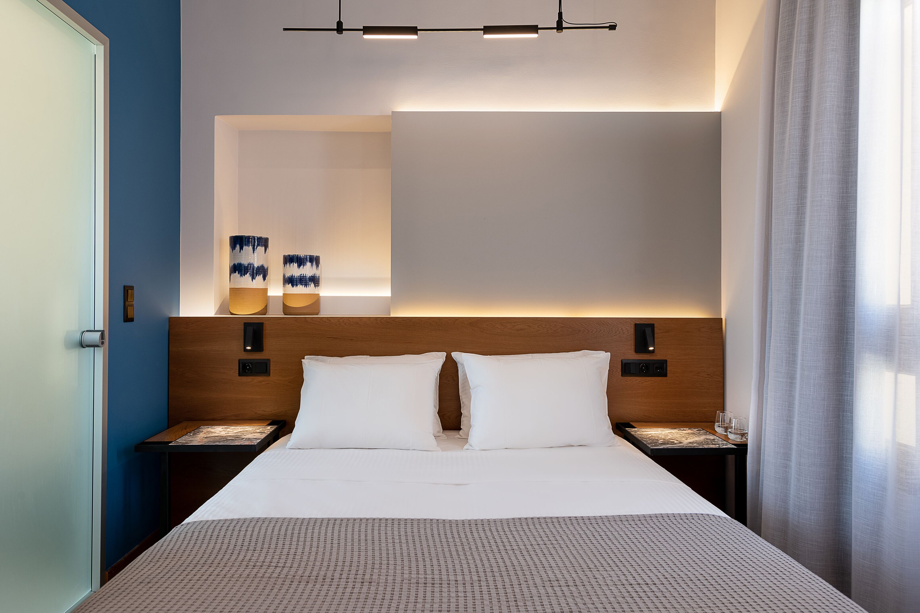 Una habitación de hotel moderna con una cama grande, mesitas de noche, un cabecero de madera y una lámpara fijada a la pared.