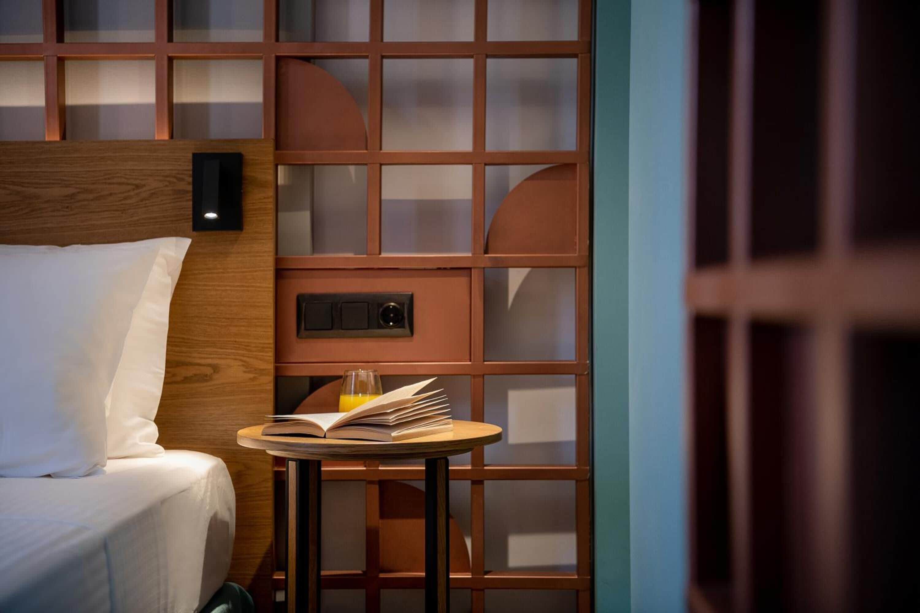 A hotel room with a bed, a small table with a book and a glass of orange juice, and a wooden headboard.