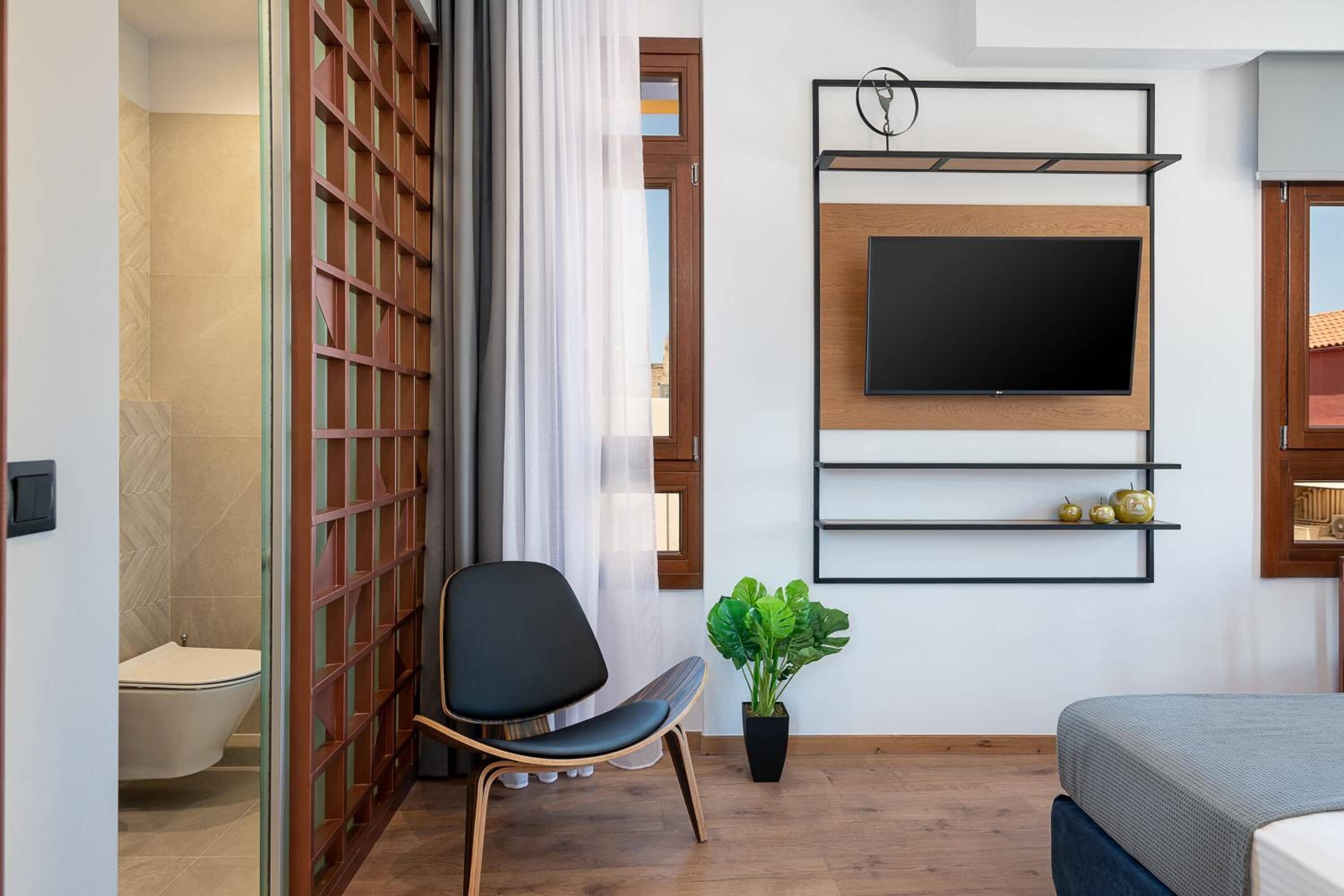A modern hotel room with a wooden floor, a black chair, a bed with a grey bedspread, a mounted TV, a potted plant, and a sliding door with a view to the outside.