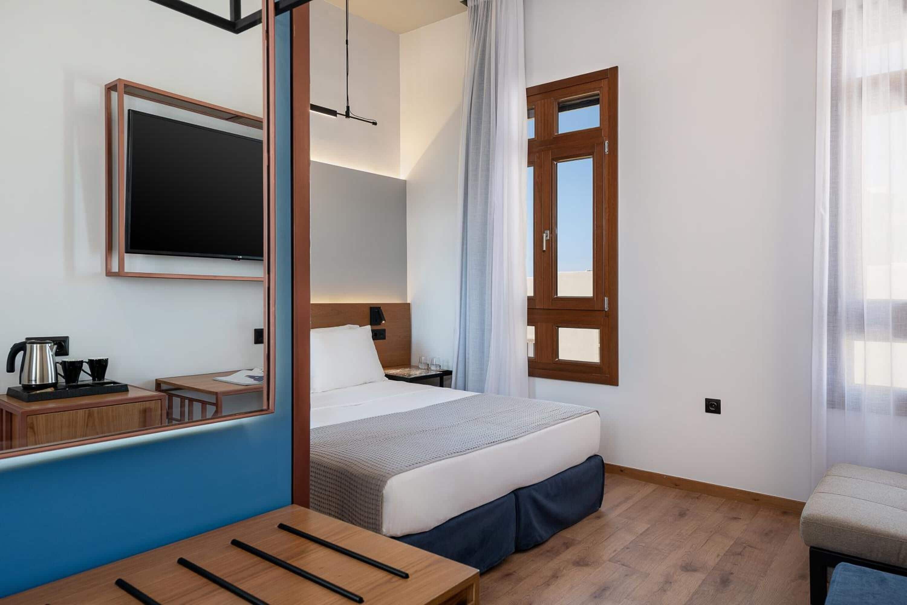 Quarto de hotel com cama, mesa de cabeceira, espelho, janela com cortinas e chão de madeira.