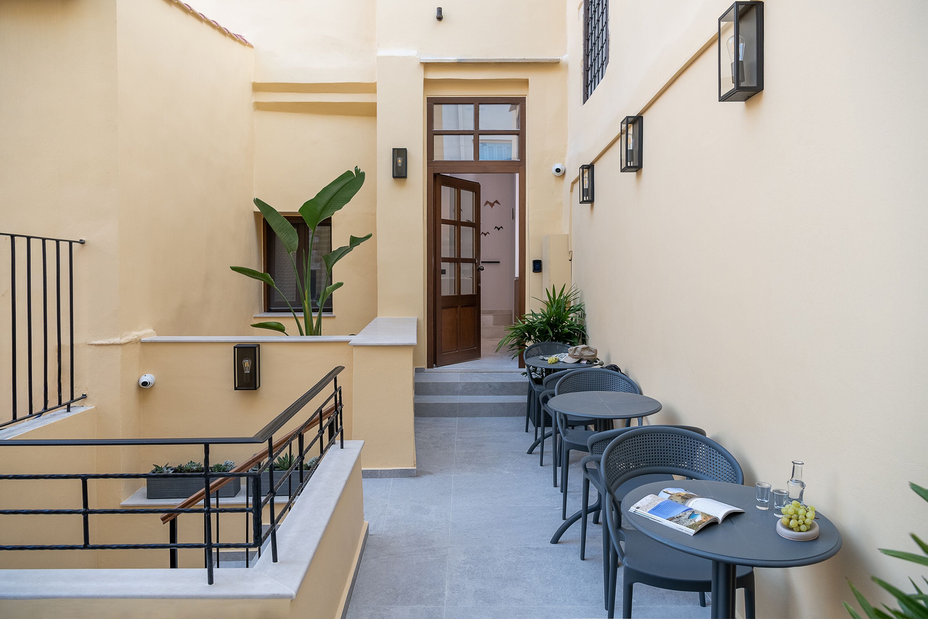 A narrow outdoor corridor with a staircase leading to an entrance, surrounded by potted plants and outdoor seating with tables and chairs.