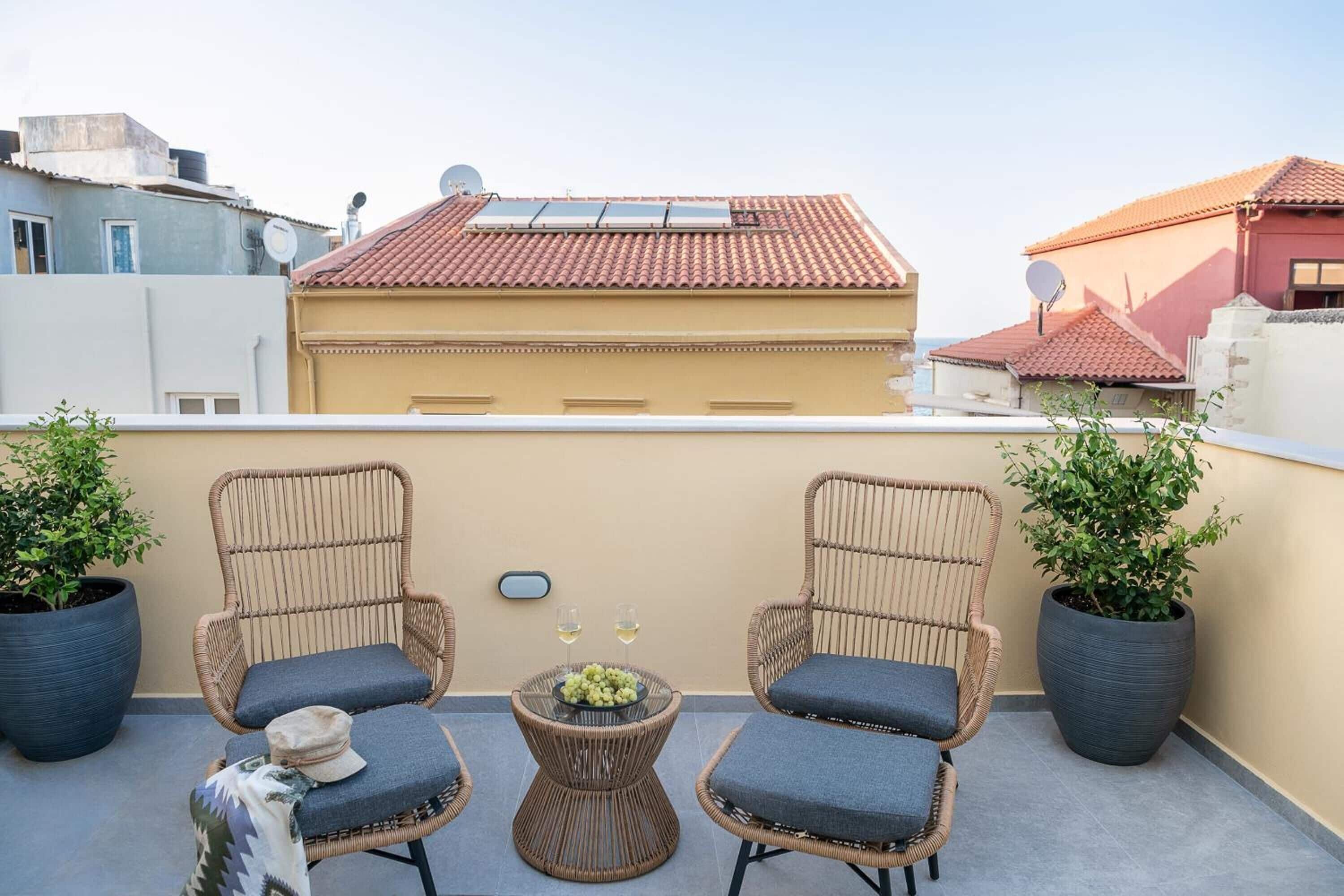 A rooftop terrace with wicker chairs, a small table with a bowl of fruit and glasses, and potted plants.