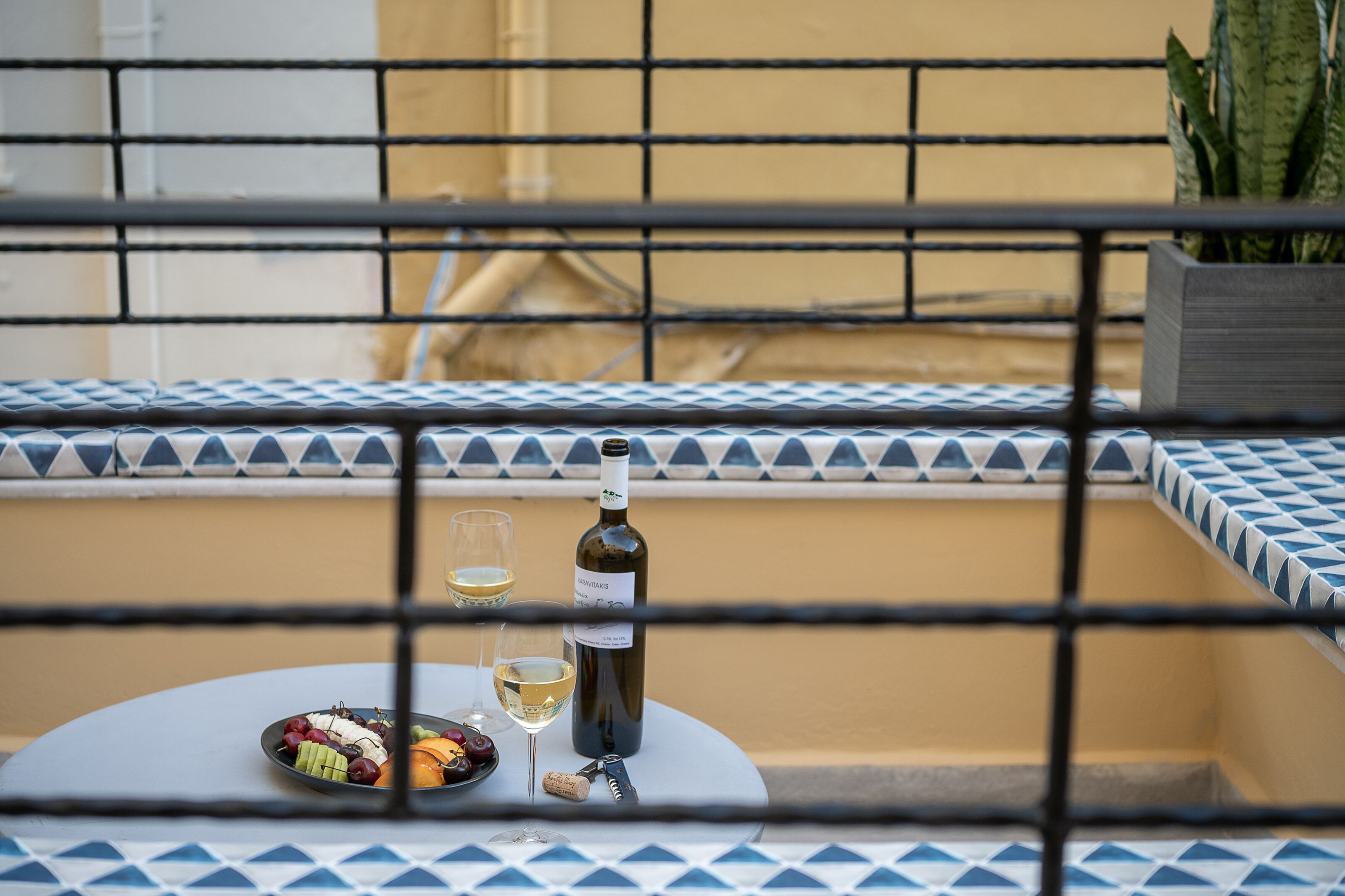 Ein Tisch mit einer Weinflasche, zwei Gläsern und einer Obstplatte auf einem Balkon mit gemusterten Kissen.