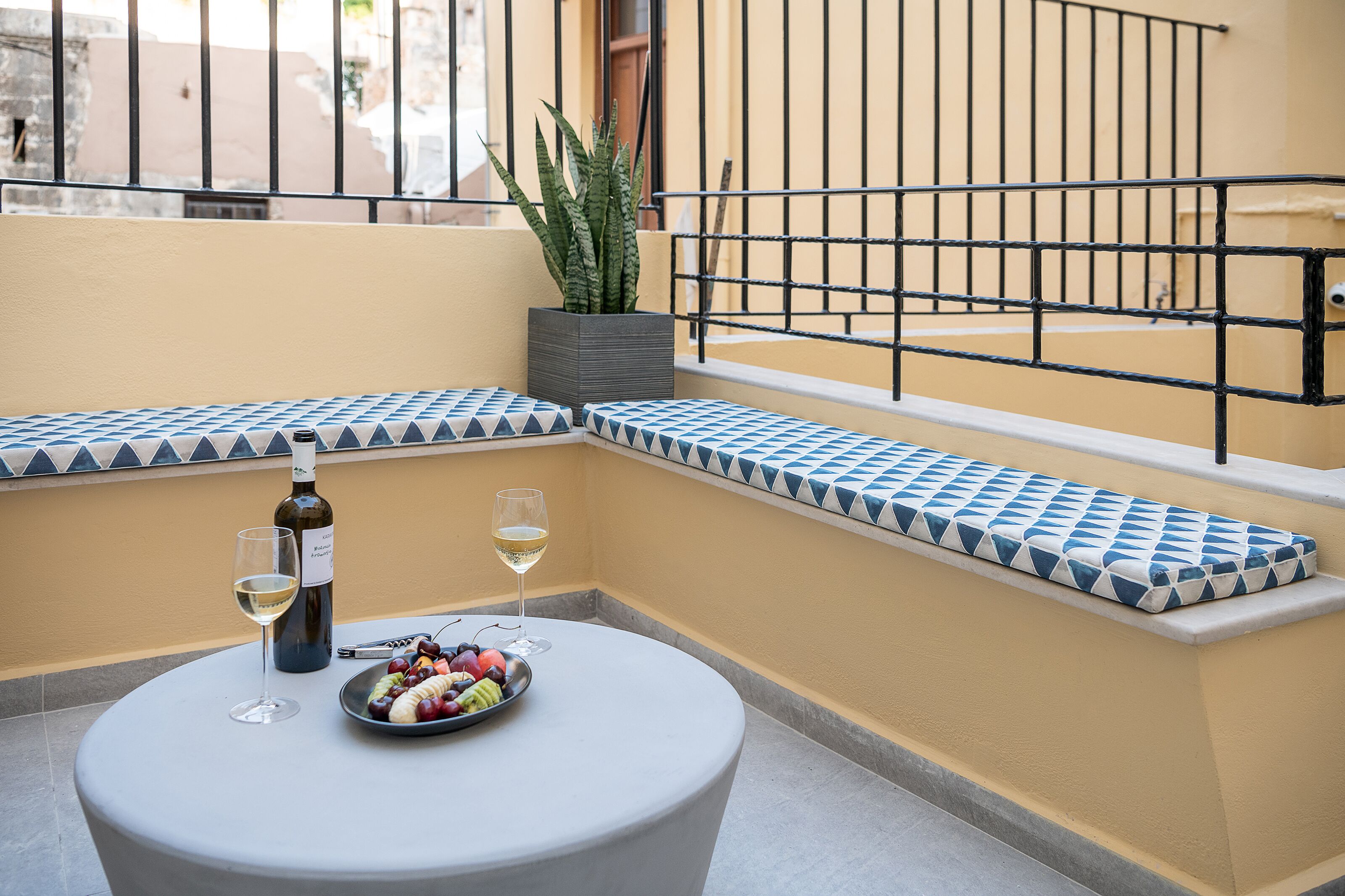 Een ronde tafel met een fles wijn, een glas en een schaal fruit op een balkon met een betegelde bank en een bloempot.