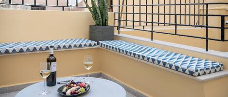 Una mesa redonda con una botella de vino, una copa y un bol de fruta en un balcón con un banco de azulejos y una planta en maceta.