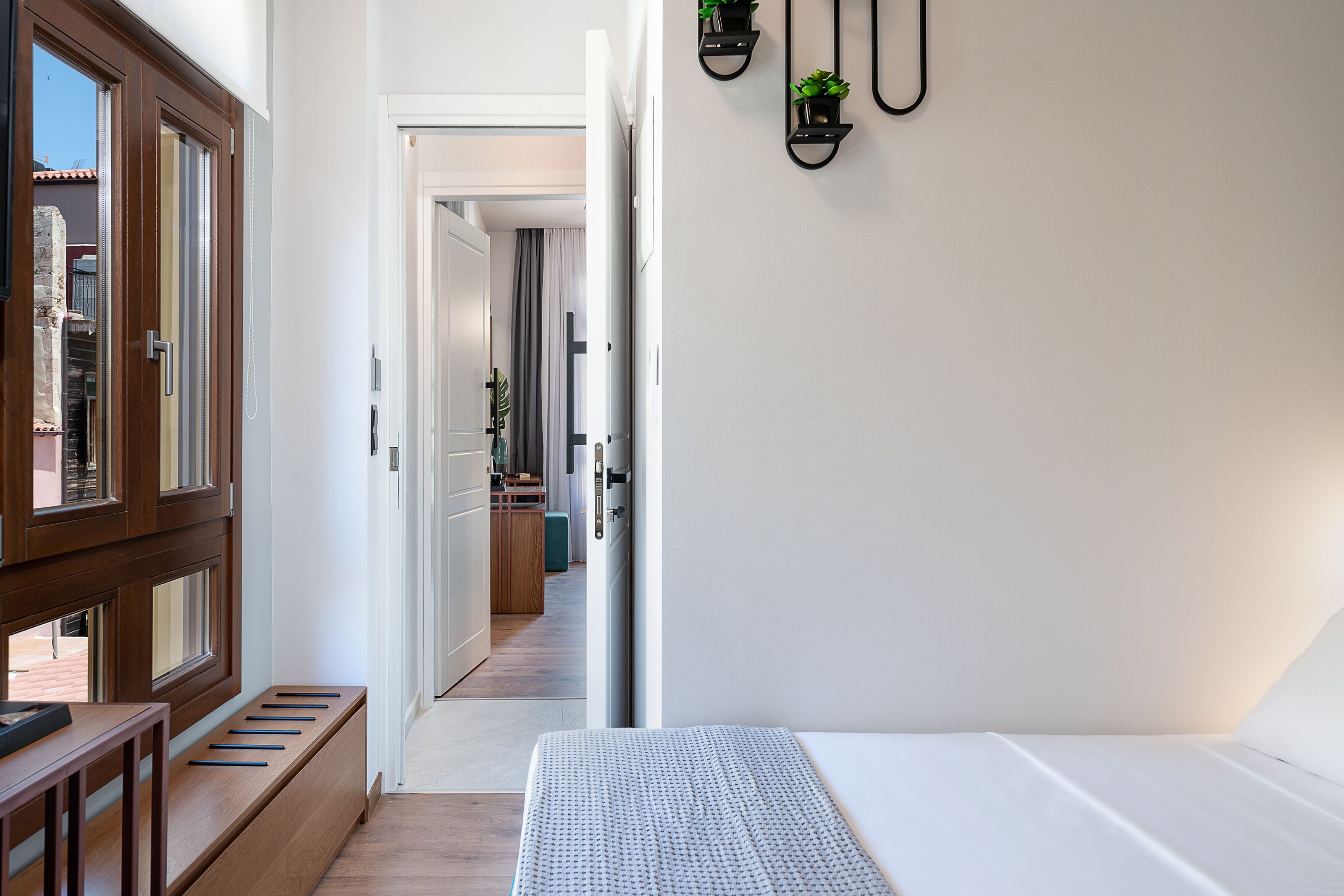 A modern bedroom with a wooden bench, a bed, and a view of the hallway leading to a kitchen.