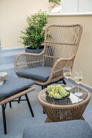 Una silla de mimbre con cojines, una mesita con un cuenco de uvas y dos copas de vino, y una planta en maceta al fondo.