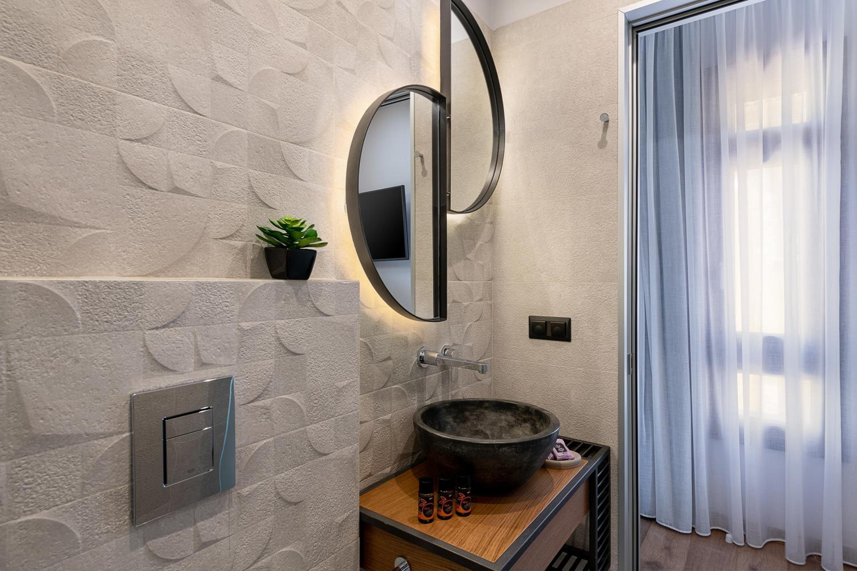 A bathroom with a stone-textured wall, a round mirror, a wooden vanity, a black vessel sink, and a small potted plant.