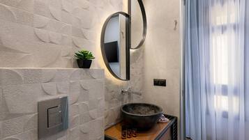 Un baño con pared de textura de piedra, un espejo redondo, un tocador de madera, un lavabo de tipo cuenco negro y una pequeña planta en maceta.