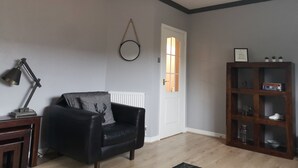 A living room with a black leather sofa, a wooden side table, a floor lamp, a wooden bookcase, and a white door with a window. - Marina Apartment by Switchback Stays (Penarth)