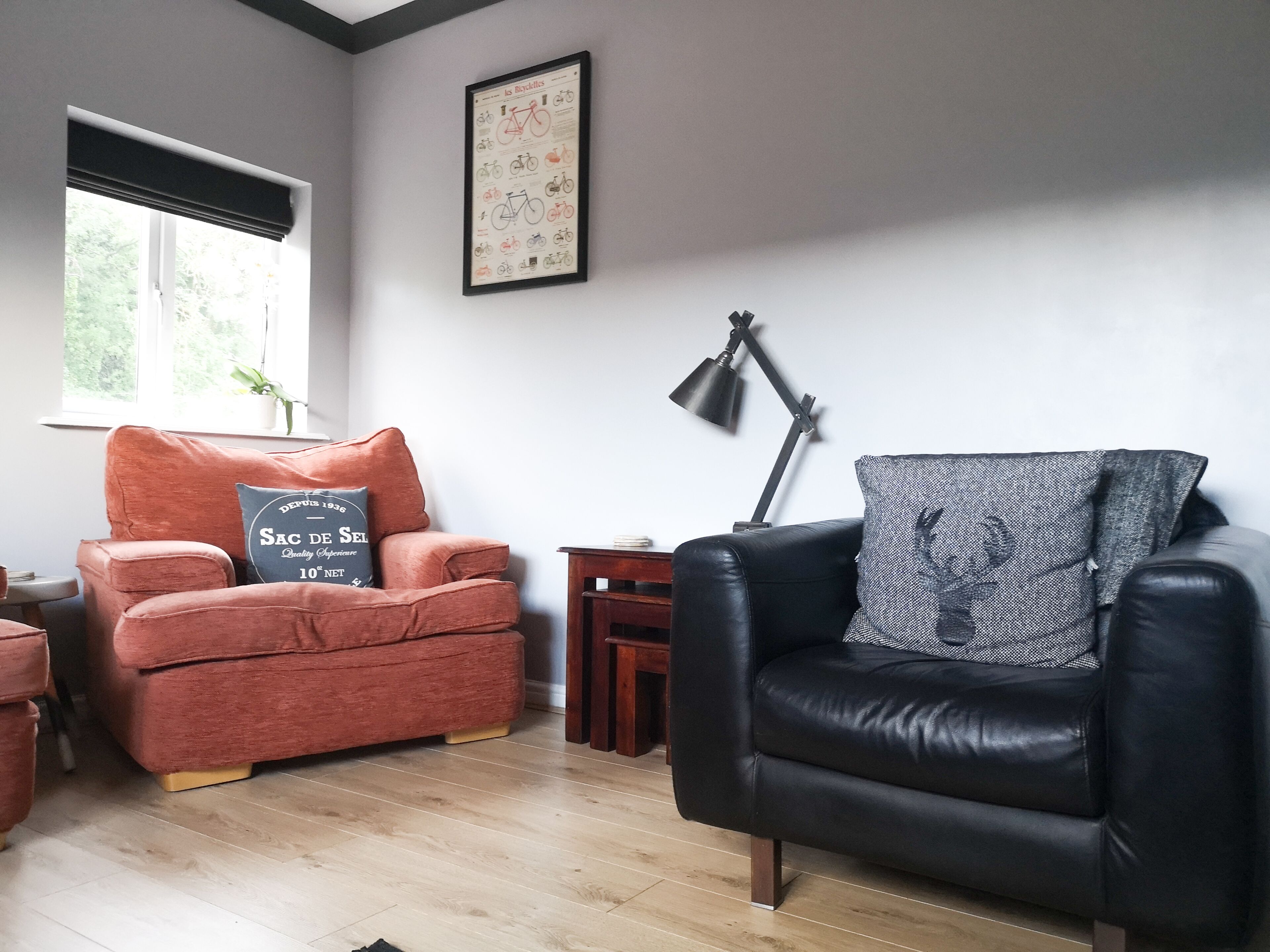 A living room with a red armchair and a black leather armchair, a framed picture on the wall, a floor lamp, and a window with a view of trees.