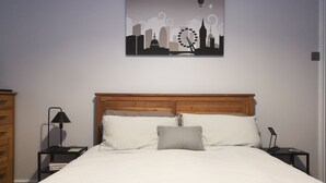 A neatly made bed with a wooden headboard, a bedside table with a lamp, and a wall-mounted artwork depicting a cityscape with a Ferris wheel. - Marina Apartment by Switchback Stays (Penarth)
