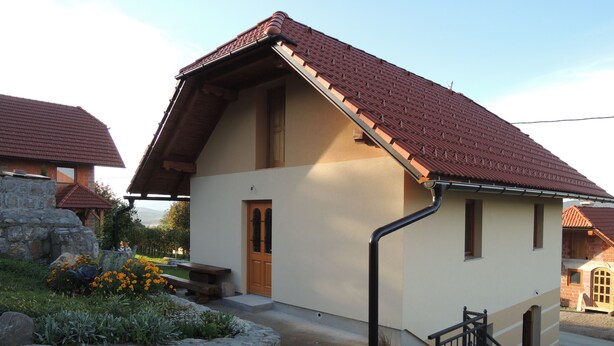 A two-story house with a sloped red-tiled roof, a stone wall, and a small garden with flowers.