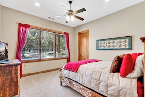 Une chambre à coucher avec un lit, un ventilateur de plafond, une fenêtre avec des rideaux, une porte en bois et un tableau encadré au mur.