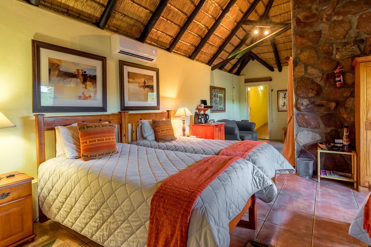 A room with a bed, a wooden nightstand, a lamp, framed artwork, a stone wall, and a wooden door.