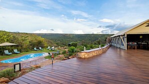Una terraza de madera con piscina, mobiliario exterior y una zona de comedor con vistas a las montañas.