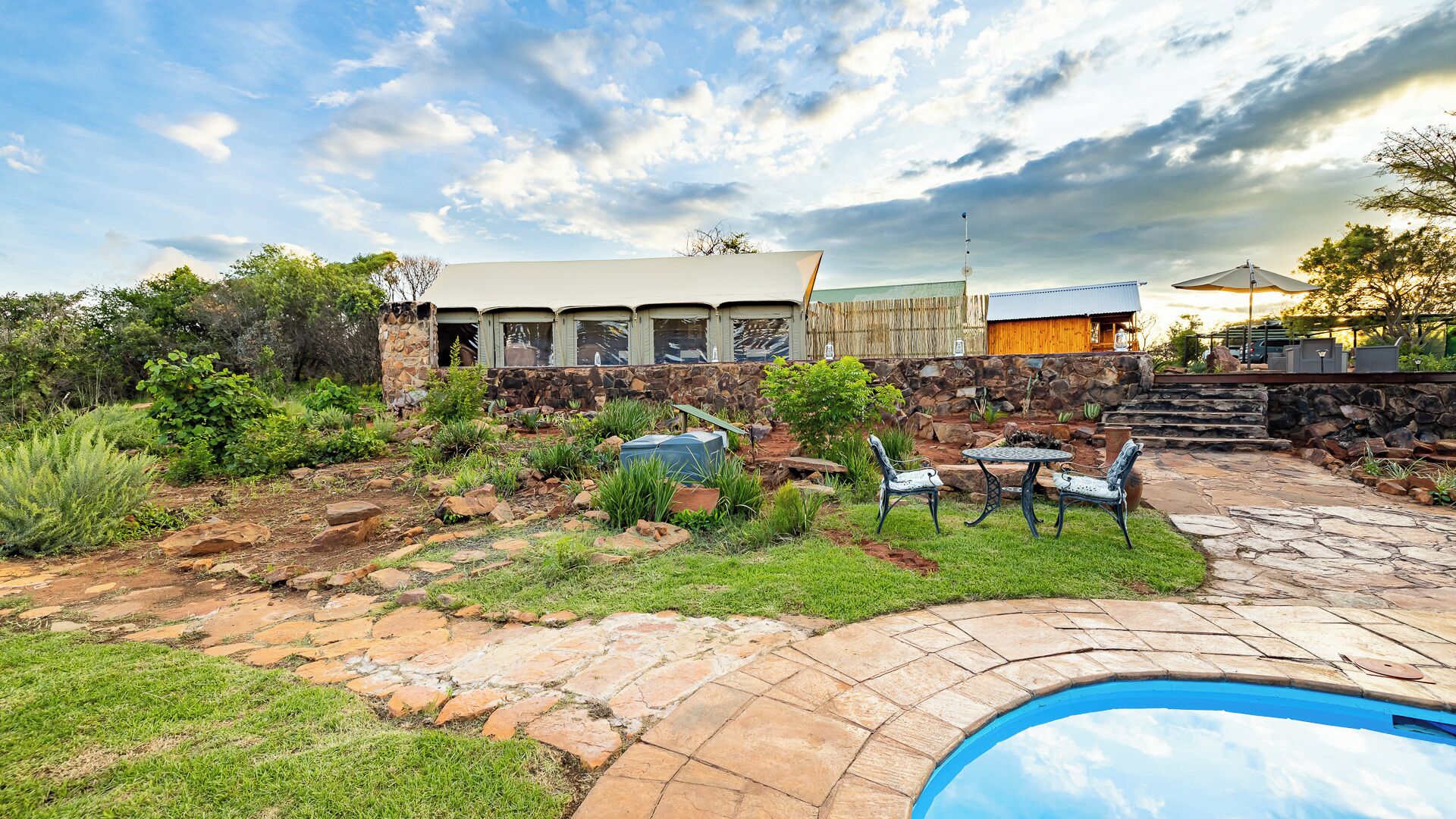 Un área de piscina con baldosas de piedra, una zona de barbacoa y un muro de piedra con una estructura de madera al fondo.