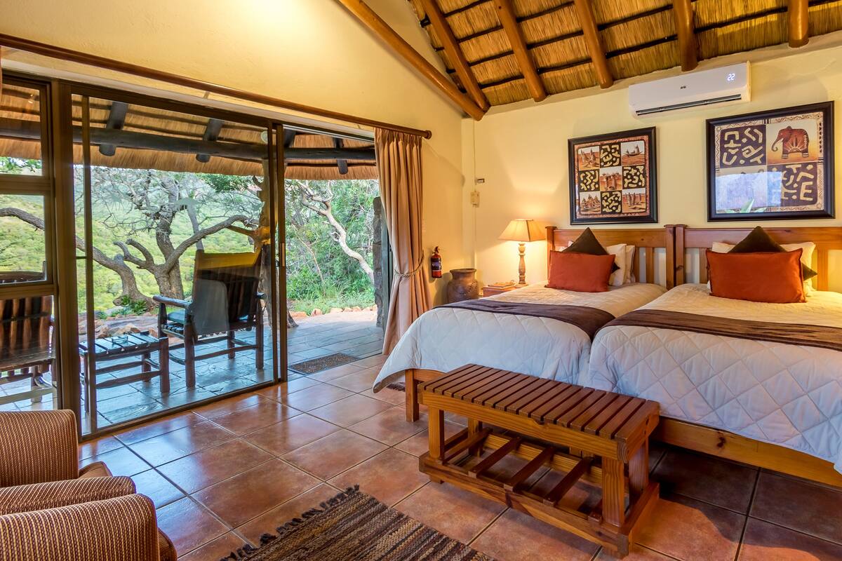A room with two beds, a wooden bench, a table, and a view of the outdoors.