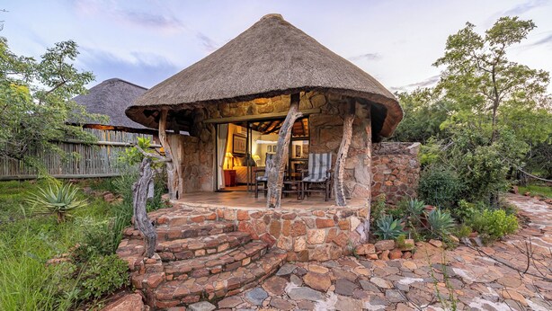 Una estructura de piedra con techo de paja, una escalera de piedra que conduce a una entrada y vista del interior con muebles y una lámpara.