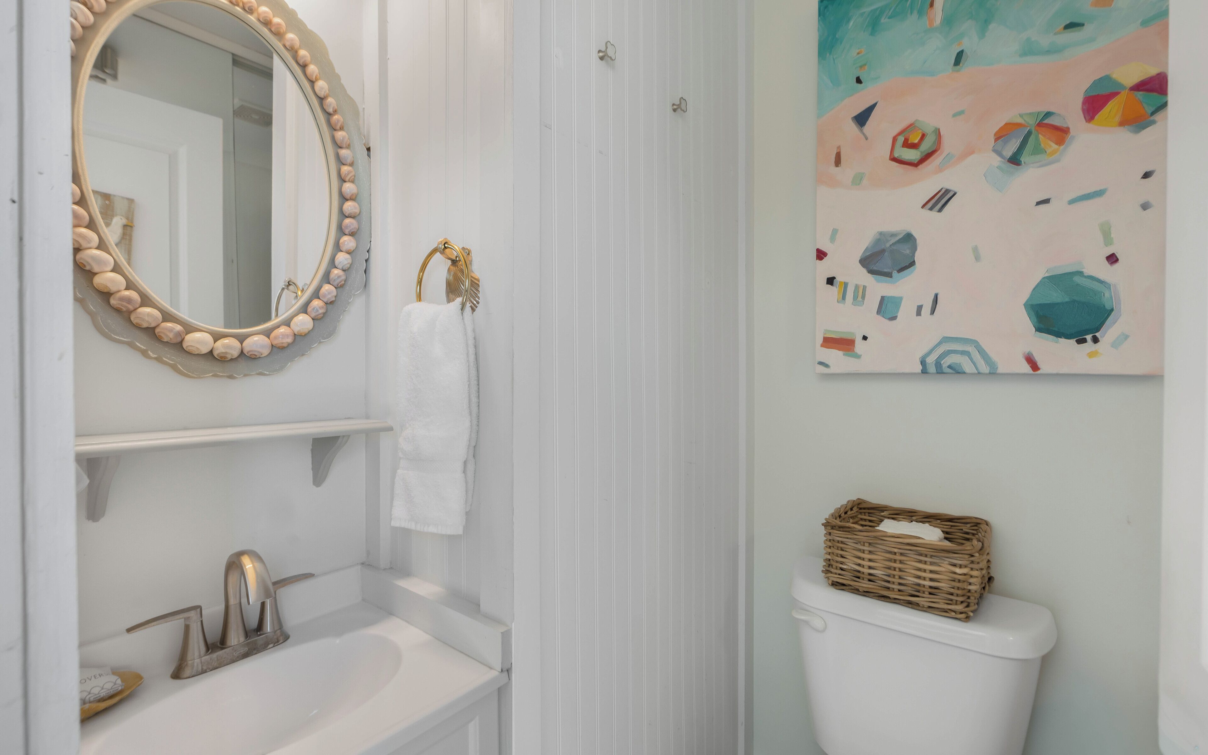 Un baño con un lavabo blanco, un grifo dorado, un espejo redondo con detalle de cuentas, una toalla blanca, un inodoro blanco y un cuadro abstracto de colores en la pared.