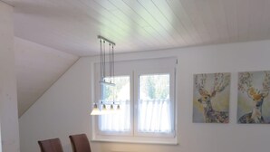 Un comedor con una mesa, sillas y un banco. Hay dos ventanas con cortinas y un techo con vigas de madera.