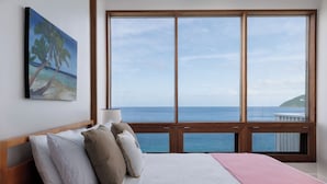 A bedroom with a large window overlooking the sea, a bed with pillows, and a painting of a beach scene. - CLIFF HOUSE ONE - SUMMER DISCOUNT, 2 min walk to 2 beaches, surf & restaurants (West End)