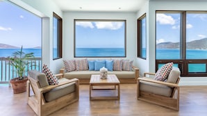 A spacious living area with a large window overlooking the sea, a sofa set, and wooden furniture. - CLIFF HOUSE ONE - SUMMER DISCOUNT, 2 min walk to 2 beaches, surf & restaurants (West End)