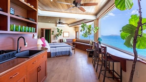 A spacious room with a wooden ceiling, a large window with a view of the ocean, a kitchenette with a sink and cabinets, and a seating area with a sofa and chairs. - CLIFF HOUSE ONE - SUMMER DISCOUNT, 2 min walk to 2 beaches, surf & restaurants (West End)