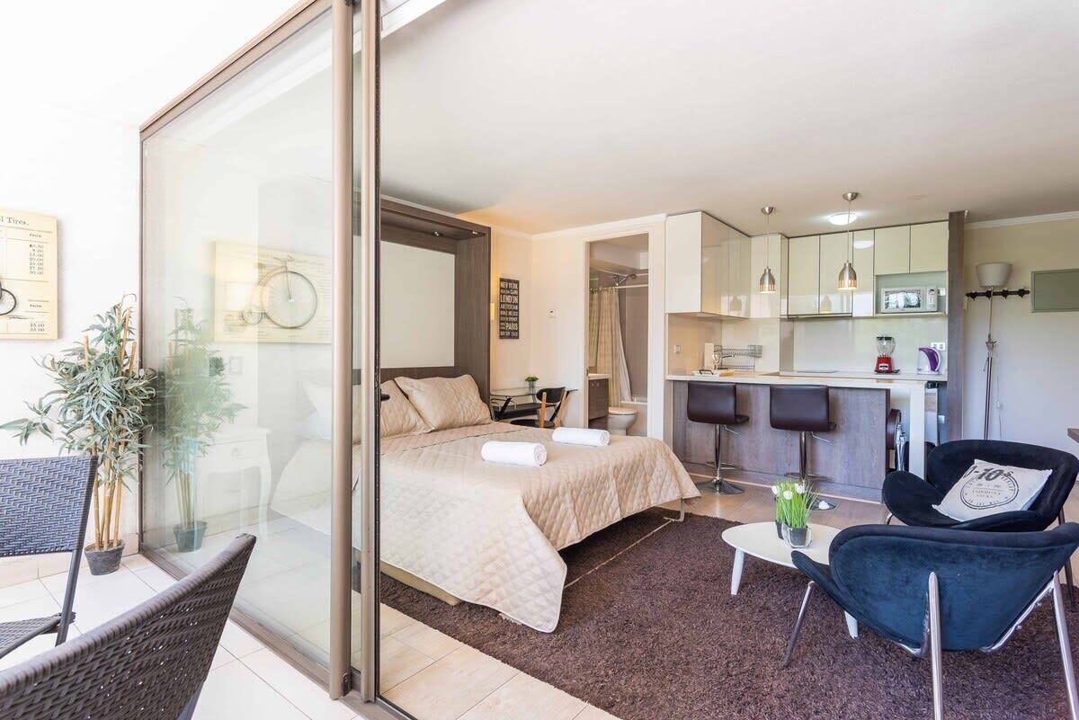 A modern hotel room with a bed, a small table, a chair, a kitchen area, and a balcony with a chair and a plant.