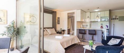 A modern hotel room with a bed, a small table, a chair, a kitchen area, and a balcony with a chair and a plant.