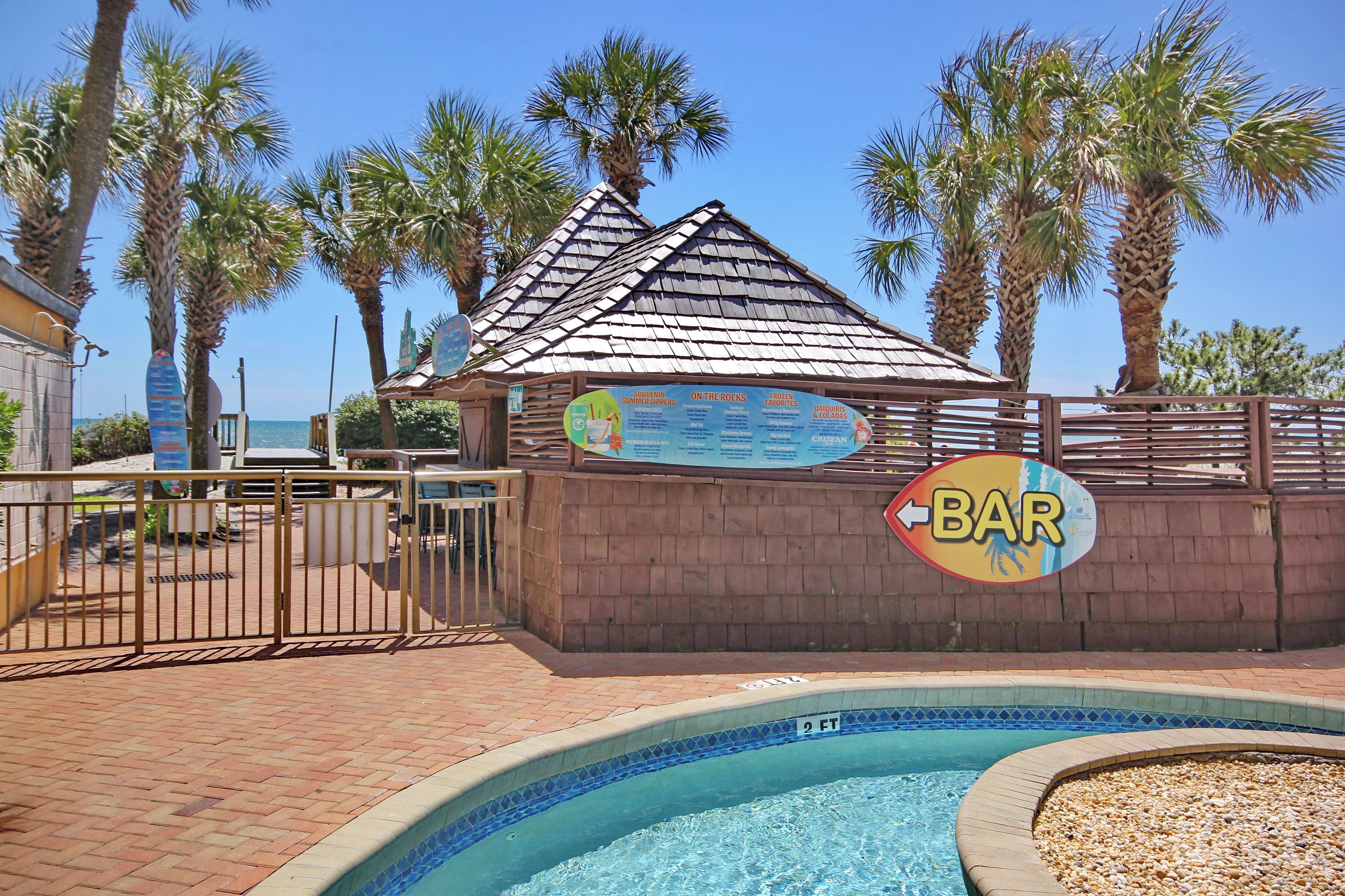 A pool area with a bar sign, a wooden structure with a shingled roof, and palm trees.