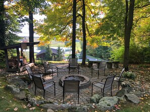 An outdoor seating area with stone benches and a fire pit, surrounded by trees and a view of a lake and buildings. - Sojourner's Lodge & Log Cabin Suites (Dundee)