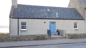 A small, single-story building with a blue door and two windows, a stone wall, and a paved entrance. - Seaview Cottage (Dundee)