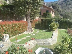 Una casa con un sendero de piedra que conduce a una zona con césped, rodeada de diversas plantas y una montaña al fondo.