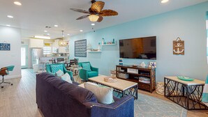 Een moderne woonkamer met een donkerblauwe bank, teal fauteuils, een houten salontafel en een plafondventilator. Op de achtergrond is een keuken met witte kastjes en een eethoek zichtbaar.