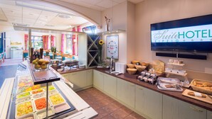 Zona de bufé del hotel con diversas opciones de comida, una pantalla mostrando el nombre del hotel y una vista del comedor al fondo.