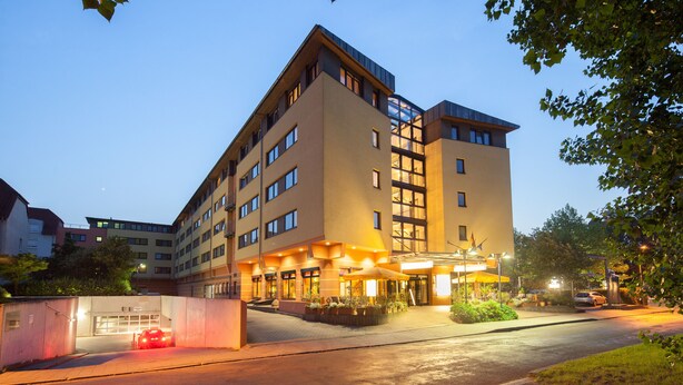 Un hotel de varios pisos con diseño moderno, que cuenta con una entrada destacada con zona cubierta y asientos al aire libre.