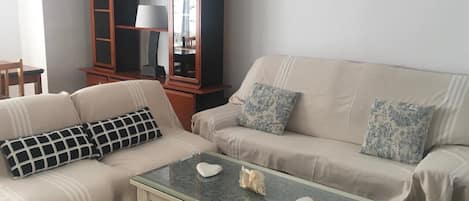 A living room with a beige sofa, a glass coffee table, and a wooden cabinet with shelves.