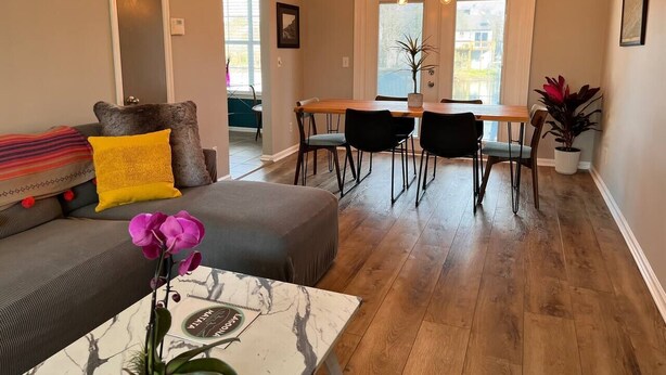 Un salon comprenant un canapé, un pouf et une table basse, un coin repas avec une table et des chaises, et un couloir menant à une autre pièce.
