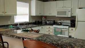 A kitchen with white cabinets, a microwave, a stove, and a refrigerator. There are two bar stools and a window with blinds.