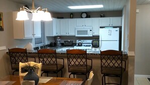 A dining area with a wooden table and chairs, a chandelier, and a kitchen in the background with white cabinets and appliances.