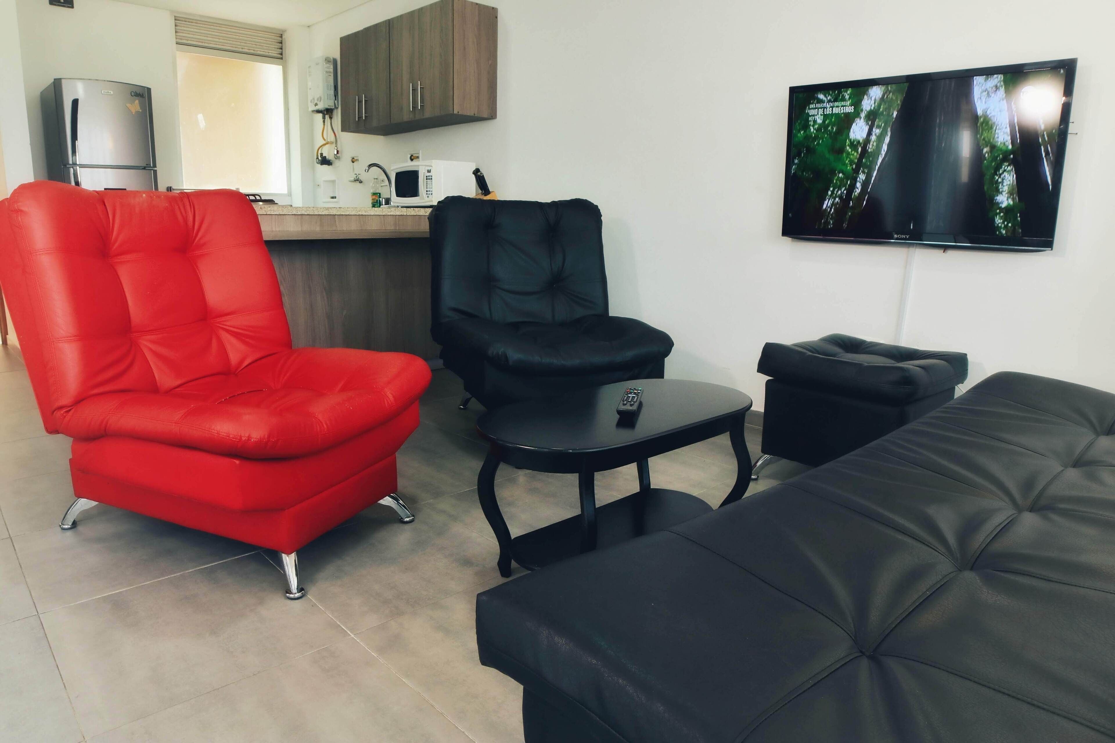 A living area with a red chair, a black chair, a black ottoman, and a black sofa. A flat-screen TV is mounted on the wall.