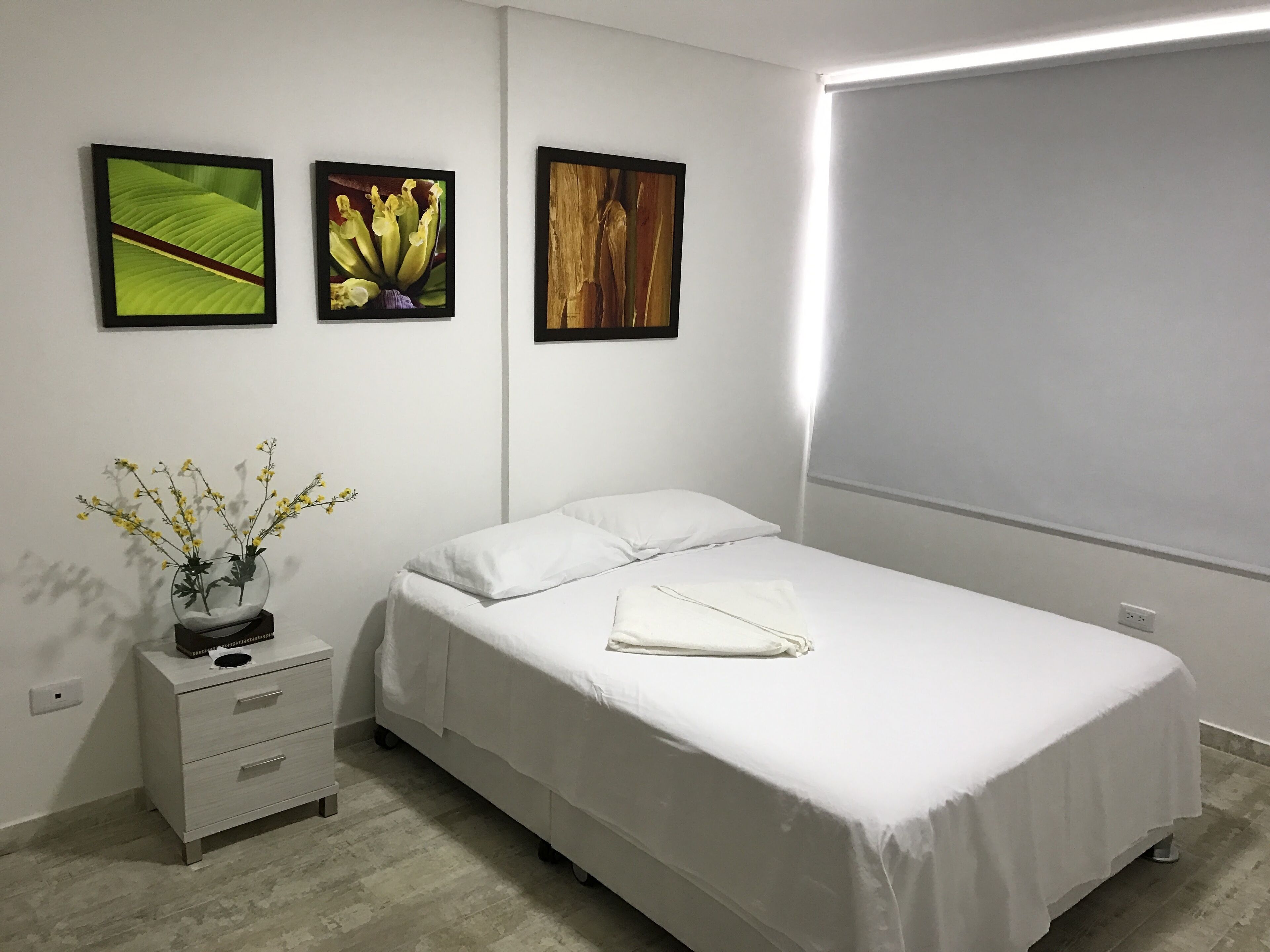 A neatly made bed with white linens, a small bedside table with a vase of flowers, and three framed pictures on the wall.