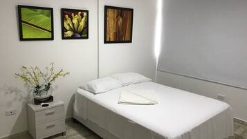 A neatly made bed with white linens, a small bedside table with a vase of flowers, and three framed pictures on the wall.