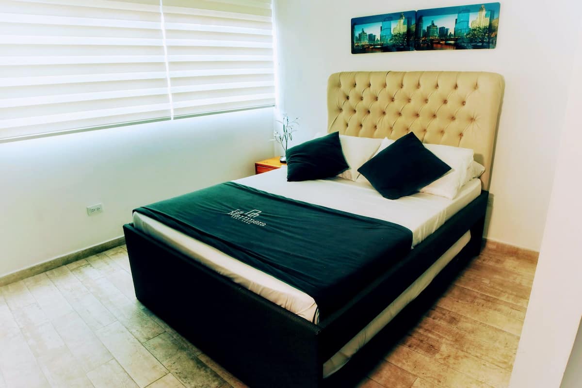 Un dormitorio con cama, mesita de noche, ventana con persianas y un cuadro de paisaje urbano en la pared.