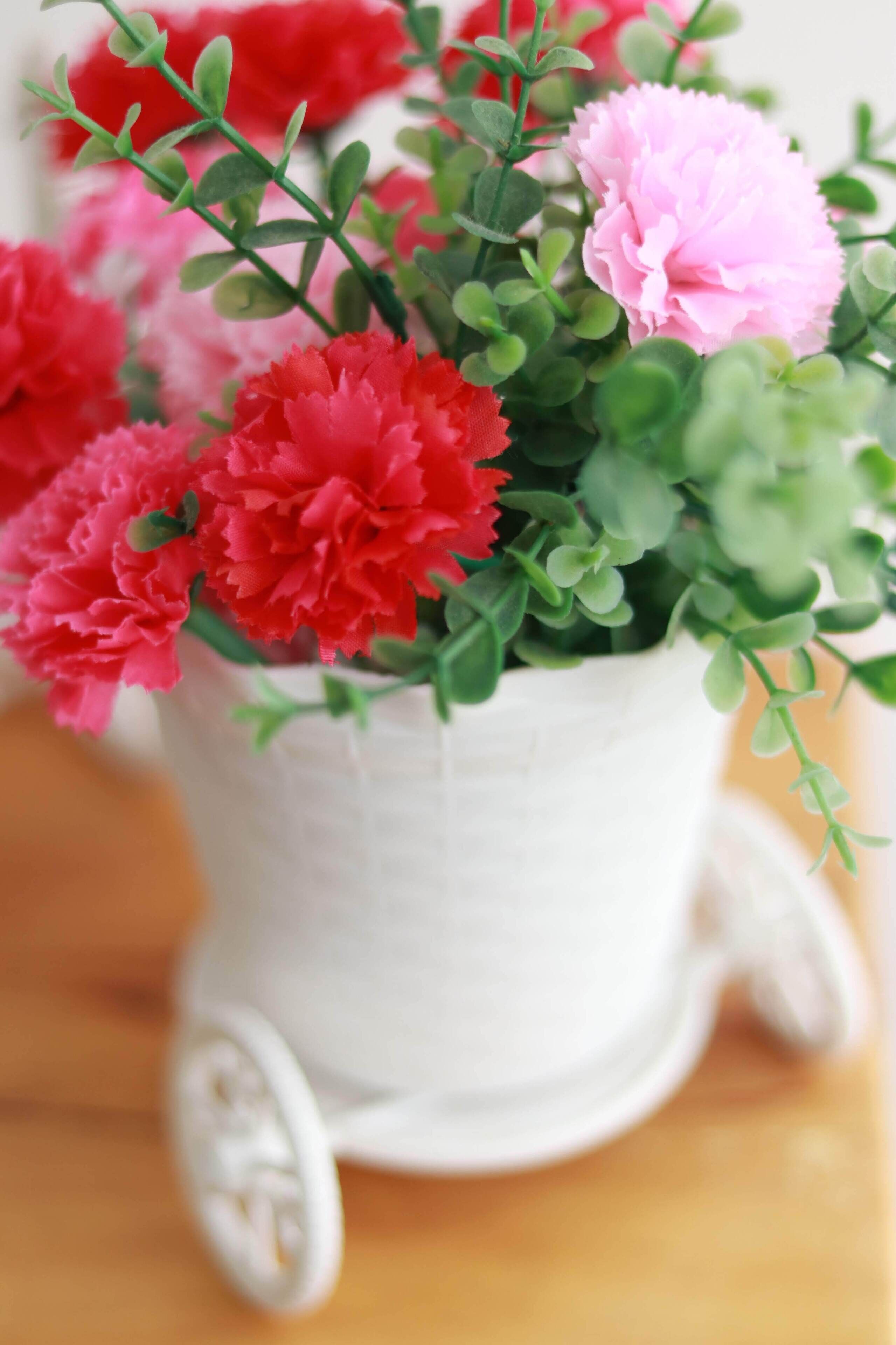 Una maceta de cerámica blanca con asa, llena de flores rosadas y rojas y follaje verde.