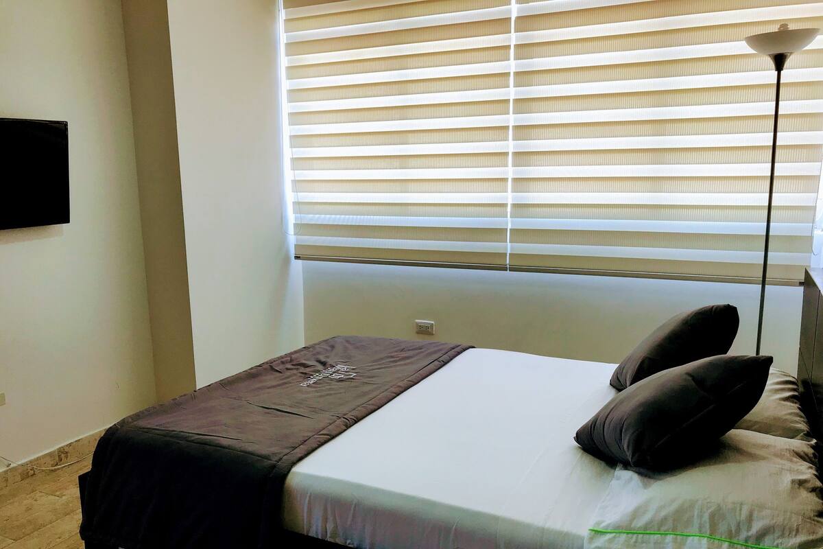 Un dormitorio con cama, ventana con persianas, lámpara de pie y televisor montado en la pared.