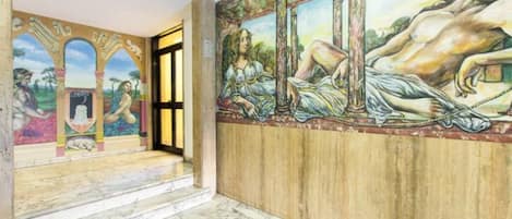 Un pasillo con un mural que representa una escena clásica, suelo de mármol y paneles de madera en la pared.